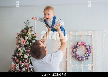 Junger Vater wirft Baby Sohn in hellen weißen Raum mit Weihnachten Interieur zu Hause. Familie, Urlaub, Elternschaft, Spiel, Kindheit und Stockfoto