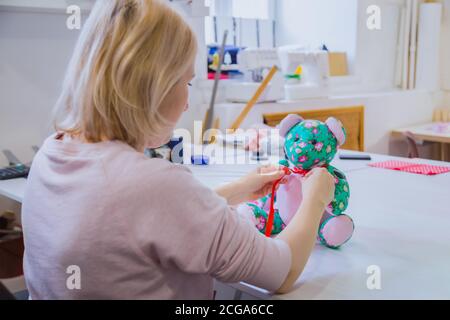 Schneiderin, Spielzeugmacher sitzt und Binde Schleife am Hals des Teddybären in der Nähwerkstatt. Handgefertigtes, handwerkliches und Spielzeug-Konzept. Nahaufnahme Stockfoto