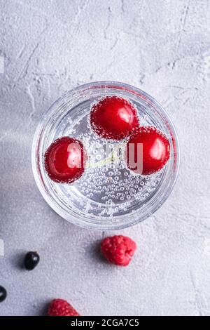 Frisches kaltes Mineralwasser Getränk mit Kirsche, Himbeere und Johannisbeere in transparentem Glas auf Steinbeton Hintergrund, sommerliche Diät Getränk Stockfoto
