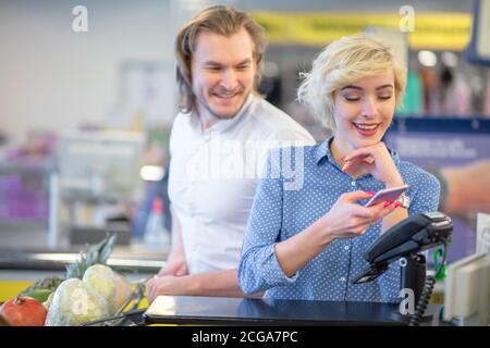 Junge Pärchen, die im Einkaufszentrum Waren kaufen, blonde hübsche Frau zeigt ihre Coupons an der Kasse und bekommt einen Rabatt, während sie mit Kreditkarte bezahlt! Stockfoto