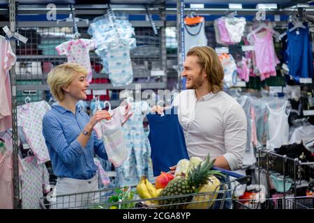 Ein kleiner Elternteil wählt Kleidung für Baby in einem Kindergeschäft. Blonde attraktive Mutter und Vater in der Mall. Konzept der Einkaufsbekleidung. Stockfoto