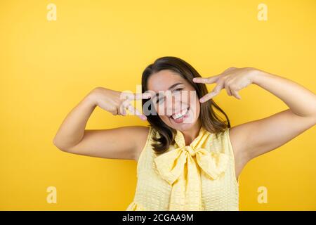 Junge schöne Frau über isolierten gelben Hintergrund tun Frieden Symbol mit Fingern über Gesicht, lächelnd fröhlich zeigt Sieg Stockfoto