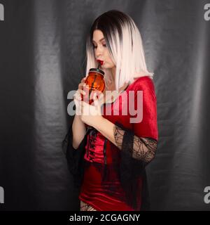 Junge schöne blonde Frau in einem roten Vampir Kleid hält Ein orangefarbenes Glas in Form eines Totenschädels Ihre Hände Stockfoto