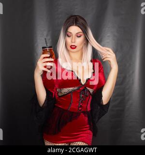 Junge schöne blonde Frau in einem roten Vampir Kleid hält Ein orangefarbenes Glas in Form eines Totenschädels Ihre Hände Stockfoto