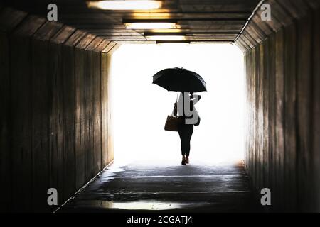 Hochgesättigte Silhouette einer selbstbewussten Frau, die einen Regenschirm hält, der aus einem Tunnel herauskommt. Bei nassem Wetter arbeiten Stadt chic Lifestyle. Wettervorhersage, Regen Stockfoto