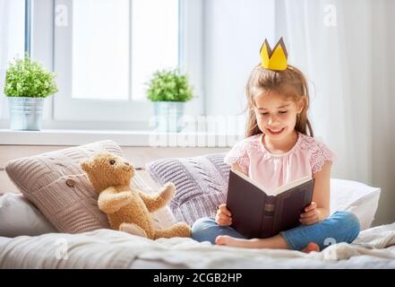 Süßes kleines Kind Mädchen das Lesen eines Buches im Schlafzimmer. Kind mit Krone auf dem Bett zu sitzen in der Nähe der Fenster. Stockfoto