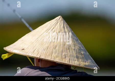 Ein asiatischer Mann, der einen traditionellen Straw Bamboo Coolie Hut trägt, steht an den hölzernen Barrieren einer Brücke an einem See und fischt mit einer Angelrute. Stockfoto