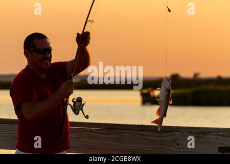 Eastern Neck Island, MD, USA 09/06/2020: Ein junger hispanic-Mann mit rotem T-Shirt und Sonnenbrille hat gerade einen Katzenfisch mit seiner Angelrute gefangen. Er Stockfoto