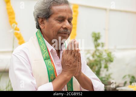 Älterer Mann in geschlossenen Augen bietet Gebet durch Namaste Geste beten zu gott zu Hause in traditioneller Kleidung während Ritualen. Stockfoto