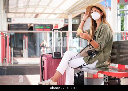 Die junge Frau trägt eine Gesichtsmaske und einen Hut und benutzt ihr Smartphone, während sie am Bahnhof auf den Zug wartet. Reisekonzept. Stockfoto