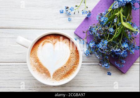 Blaue Vergissmeinnicht-Blumen, Notizbuch und eine Tasse heißen Kaffee. Das Konzept der Feiertage und Guten Morgen Wünsche. Stockfoto