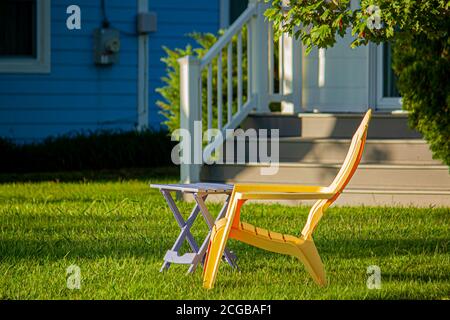 Nahaufnahme Bild von einem einfachen gelben Plastikstuhl und einem klappbaren Tisch im Freien auf dem Vorgarten eines Hauses an einem sonnigen Tag platziert. Ein einfacher Garten konz Stockfoto