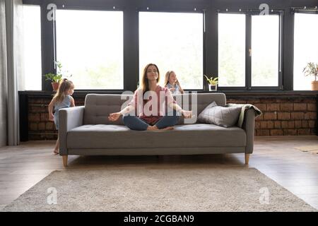 Ruhige Frau konzentriert sich auf Yoga-Übungen auf Couch zu Hause, während zwei laute Kinder lachen, laufen Stockfoto