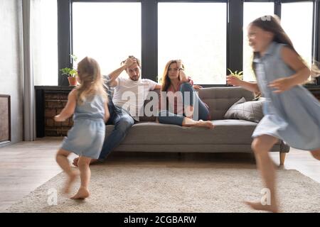 Müde Mutter und Vater sitzen auf der Couch fühlt sich genervt erschöpft Während die Töchter laut schreien, laufen sie um das Sofa herum Stockfoto