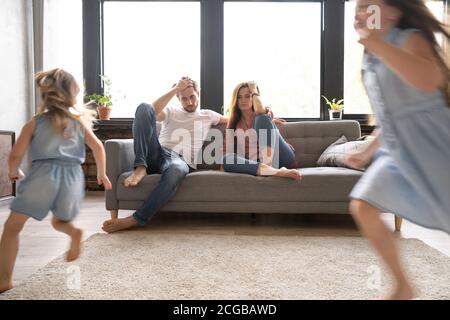 Müde Mutter und Vater sitzen auf der Couch fühlt sich genervt erschöpft Während die Töchter laut schreien, laufen sie um das Sofa herum Stockfoto