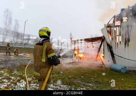 Norwegischer Feuerwehrmann von hinten versucht, Flammen auf Haus in Brand mit riesigen Flammen und Rauch, im Winter mit Schnee zu löschen. Stockfoto