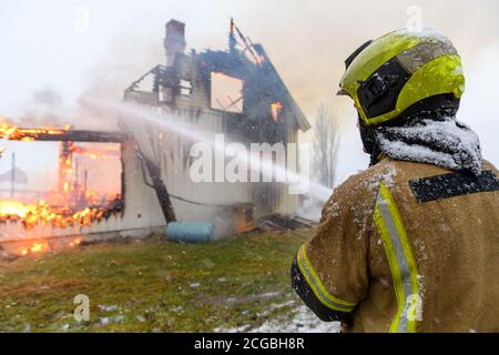 Norwegischer Feuerwehrmann von hinten versucht, Flammen auf Haus in Brand mit riesigen Flammen und Rauch, im Winter mit Schnee zu löschen. Stockfoto