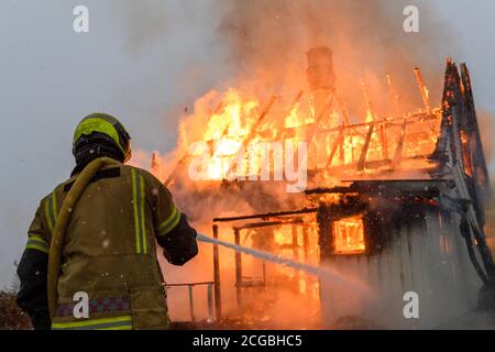 Norwegischer Feuerwehrmann von hinten versucht, Flammen auf Haus in Brand mit riesigen Flammen und Rauch, im Winter mit Schnee zu löschen. Stockfoto