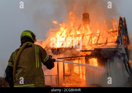 Norwegischer Feuerwehrmann von hinten versucht, Flammen auf Haus in Brand mit riesigen Flammen und Rauch, im Winter mit Schnee zu löschen. Stockfoto