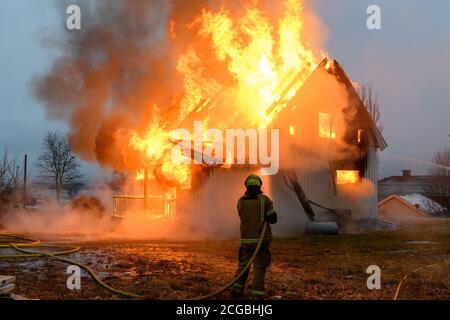 Norwegischer Feuerwehrmann von hinten versucht, Flammen auf Haus in Brand mit riesigen Flammen und Rauch, im Winter mit Schnee zu löschen. Stockfoto