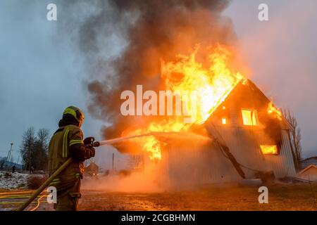Norwegischer Feuerwehrmann von hinten versucht, Flammen auf Haus in Brand mit riesigen Flammen und Rauch, im Winter mit Schnee zu löschen. Stockfoto