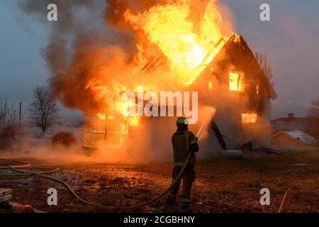 Norwegischer Feuerwehrmann von hinten versucht, Flammen auf Haus in Brand mit riesigen Flammen und Rauch, im Winter mit Schnee zu löschen. Stockfoto