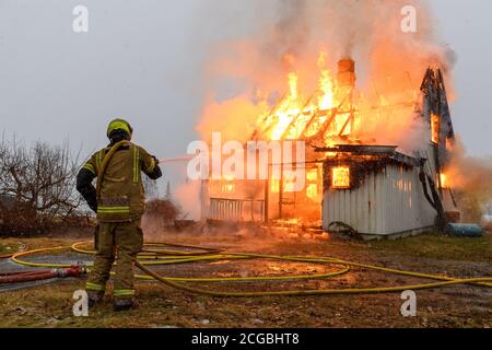 Norwegischer Feuerwehrmann von hinten versucht, Flammen auf Haus in Brand mit riesigen Flammen und Rauch, im Winter mit Schnee zu löschen. Stockfoto