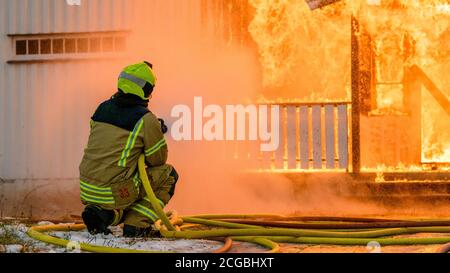 Norwegischer Feuerwehrmann von hinten versucht, Flammen auf Haus in Brand mit riesigen Flammen und Rauch, im Winter mit Schnee zu löschen. Stockfoto
