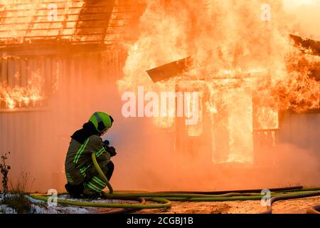 Norwegischer Feuerwehrmann von hinten versucht, Flammen auf Haus in Brand mit riesigen Flammen und Rauch, im Winter mit Schnee zu löschen. Stockfoto