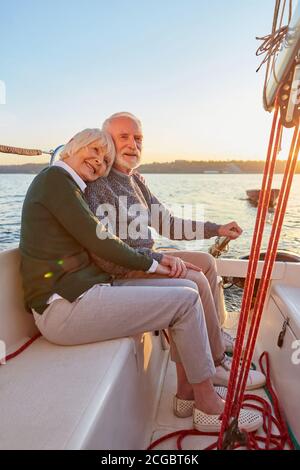 Volle Beinfreiheit von glücklichem älteren Paar, älterer Mann und Frau, die Hände halten, umarmen und Zeit zusammen verbringen, während sie auf der Seite des Yachtdecks sitzen Stockfoto