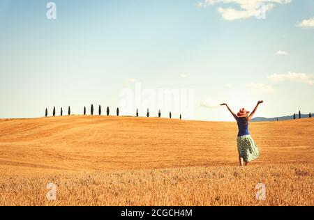 Glückliche Frau genießen die Freiheit, draußen in einem Weizenfeld. Zypressen am Horizont mit blauem Himmel und weißem Wolkenhintergrund. Stockfoto