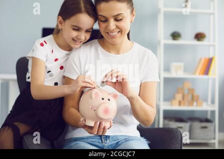 Glückliche junge Mutter und ihre schöne Tochter setzen Münzen in Sparschwein zusammen zu Hause, kopieren Raum Text Stockfoto