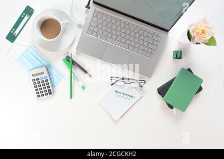 Weißer Schreibtisch mit Laptop, grüne Arbeitsmittel, chirurgische Gesichtsmaske gegen Coronavirus und covid-19 Infektion und Notizblock mit Text New Normal Stockfoto