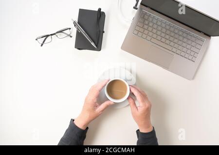 Ältere Hände halten eine Kaffeetasse auf einem weißen Schreibtisch mit Laptop, Notebook, Stift und Brille, Kopierfläche, Blick von oben Stockfoto