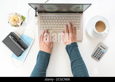 Hände einer älteren Frau, die auf einer Laptop-Tastatur tippt, weißer Schreibtisch mit Arbeitsmitteln und eine chirurgische Maske gegen Coronavirus und covid-19 inf Stockfoto