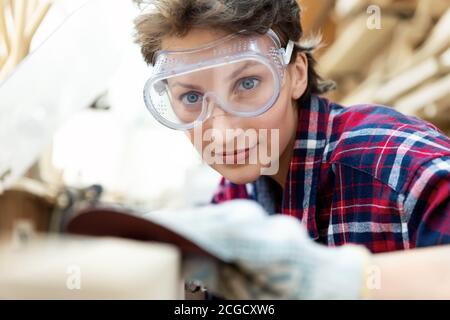 Junge professionelle schöne 30-40 weibliche Zimmermann suchen und Schleifen von rohem Holz mit Schleifpapier in Schreinerei diy Werkstatt. Weibliche Gleichberechtigung Stockfoto