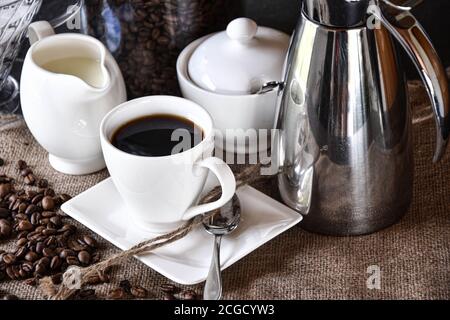 Eine Tasse Kaffee mit weißen Gerichten, ein Milchkännchen und eine Zuckerkanne auf einer Jute-Textur mit silbernen Thermoskanne serviert Kaffeekanne Stockfoto