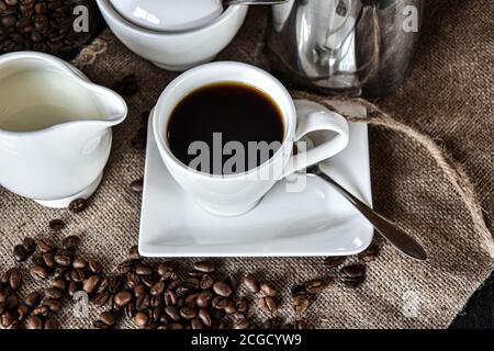 Eine Tasse Kaffee mit weißem Geschirr wie Milchkännchen, Zuckertopf und Untertasse heiß serviert auf einer Jute Textur mit Kaffeebohnen Dekoration Stockfoto