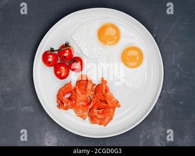 Zwei Spiegeleier Frühstück mit vier gegrillten Kirschtomaten und Lachsfisch in Scheiben auf grauem Hintergrund Draufsicht Stockfoto