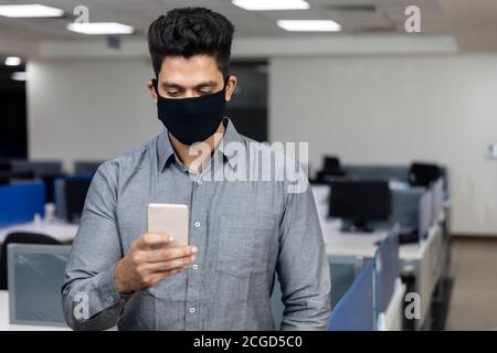 Porträt eines hübschen jungen indischen Geschäftsmann trägt Covid Schutzmaske und Überprüfung seines Telefons in einem Büro-Umgebung. Stockfoto