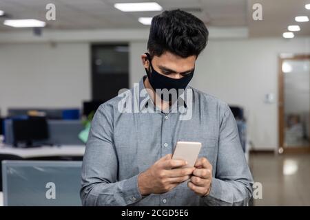 Porträt eines hübschen jungen indischen Geschäftsmann trägt Covid Schutzmaske und Überprüfung seines Telefons in einem Büro-Umgebung. Stockfoto