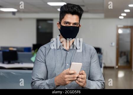 Porträt eines hübschen jungen indischen Geschäftsmann trägt Covid Schutzmaske und Überprüfung seines Telefons in einem Büro-Umgebung. Stockfoto