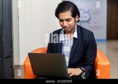 Porträt des jungen erfolgreichen indischen Geschäftsmann in dunklen Anzug sitzen auf der Couch und arbeiten auf Laptop, Corporate Environment. Stockfoto