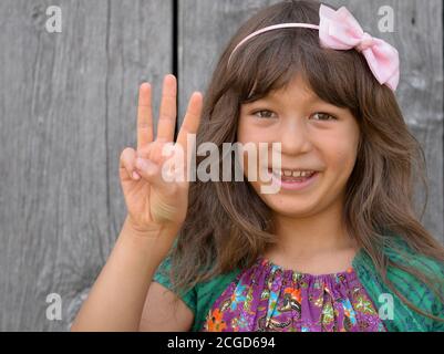 Niedliches Mischrassen-Mädchen (Ostasiatisch / Kaukasisch) zeigt mit der rechten Hand das chinesische Handzeichen für die Nummer 3 (Fotoserie: Bild-Nr. 3 von 10). Stockfoto