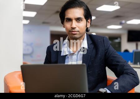 Porträt des jungen erfolgreichen indischen Geschäftsmann in dunklen Anzug sitzen auf der Couch und arbeiten auf Laptop, Corporate Environment. Stockfoto