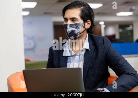 Porträt des jungen erfolgreichen indischen Geschäftsmann in dunklen Anzug tragen Covid Schutzmaske und die Arbeit an Laptop, Corporate Environment. Stockfoto