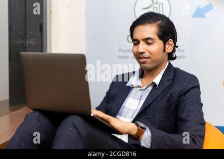 Porträt des jungen erfolgreichen indischen Geschäftsmann in dunklen Anzug sitzen und arbeiten auf Laptop, Corporate Umwelt, ausdrucksstarkes Gesicht. Stockfoto