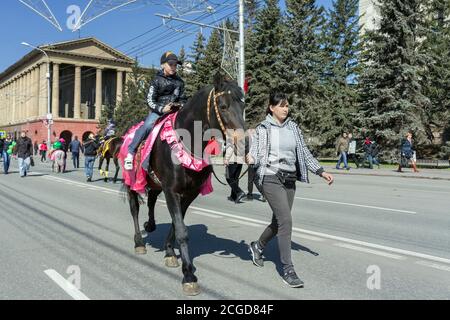 Boy reitet auf einem smarten braunen Pferd von einem Mädchen entlang der Straße von Krasnojarsk Stadt während der Feier des Siegestages 2. Weltkrieg gefahren. Stockfoto