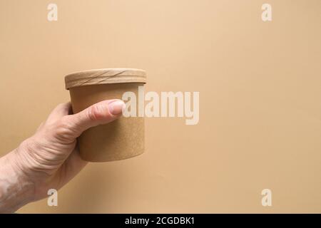 Einwegkarton Suppentasse in Womans Hand auf braunem Papierhintergrund mit Kopierplatz. Ökologisches Einzelpaket. Stockfoto