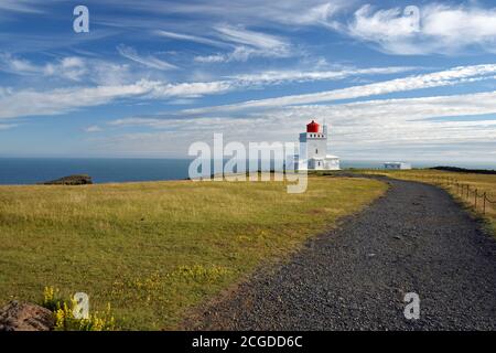 Blick auf einem vulkanischen schwarzen Steinpfad zum Leuchtturm Dyrhólaey entlang der Südküste Islands. Wispy Wolken füllen den hellen blauen Himmel. Stockfoto